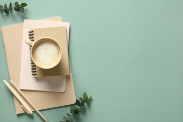 Fototapete - Stack of notebooks, cup of coffee and office supplies on a green background with eucalyptus leaves. Modern home office desk table. Flat lay, top view.