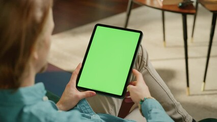 Wall Mural - Young Man Scrolling Content on Tablet Computer with Green Screen Mock Up Display. Male Relaxing at Home, Reading Social Media Posts on Mobile Device. Close Up Over the Shoulder Footage.