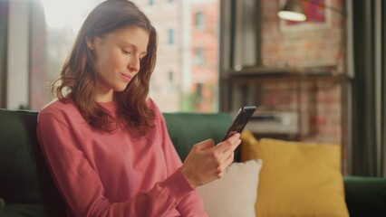 Leinwandbilder - Young Beautiful Female Sitting on a Couch in Living Room, Relaxing and Using Smartphone. Creative Girl Checking Social Media, Chatting with Friends, Browsing Internet. City View from Big Window.