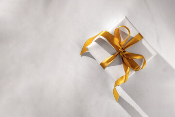 Gift box wrapped in white paper with a golden bow on festive light background. Copyspace for your text. Flat lay style. Christmas, New Year or birthday celebration concept. Web element for postcard