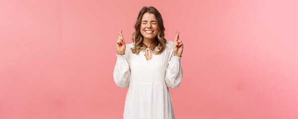 Wall Mural - Portrait of hopeful lucky cute blond girl in white dress praying, anticiapte miracle, making wish to win, cross fingers good luck, smiling and close eyes and pleading god, pink background