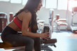 © chika_milan - Young active woman taking a pause in the gym. She is sitting, holding a water bottle with a towel on her shoulders.