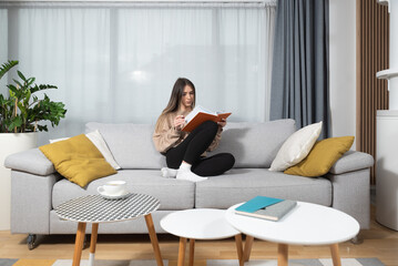 Young beautiful woman sitting on the sofa at cozy home reading the book and enjoying her free time without her husband and children with smile and happy face. Female student study and relaxing.