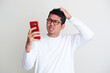 Adult Asian man scratching his head and showing confused expression while looking to his mobile phone