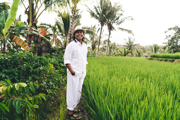 Wall Mural - Cheerful Balinese entrepreneur in hat and white wear smiling while visiting own farmland with rice fields and coffee bushes - enjoying business lifestyle at countryside of Indonesia, happiness