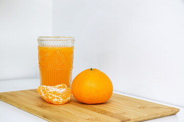 Glass of sweet and natural Mandarin orange juice, full of vitamin and nutritional goodness, besides four peeled orange pieces and a full round orange on a wooden board against white background