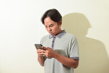 Pose Of Angry Or Annoyed Asian Man On His Smartphone In Hand. Illustration Of Indonesian Man Angry With His Cell Phone. A Man Wearing A Gray T-shirt On A White Background Isolated