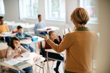 Wall Mural - Rear view of elementary school teacher points at one of her students who is raising arm to answer a question.