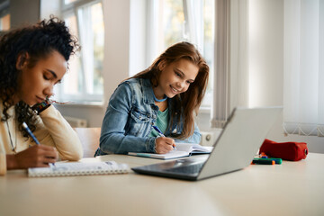 Wall Mural - Happy high school student writes while e-learning on laptop with her classmate in the classroom.