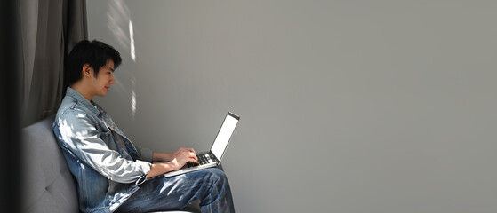 A portrait of an Asian man sitting on a couch in a living room looking at a laptop and reading something, for business, education, home and technology concept.