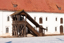 An Old Wooden Stairways To The Second Floor Outside. A Stone White Building With A Red Rusty Roof. Sunny Winter Day