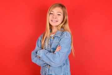 Wall Mural - blonde little kid girl wearing denim jacket over red background happy face smiling with crossed arms looking at the camera. Positive person.