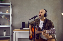 Middle Aged Male Musician Rehearsing His New Song In His Modern Home Studio. Bald Man Wearing Headphones Sitting On Chair, Playing Music On Acoustic Guitar And Singing Into Microphone