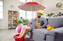 Funny Man Spends Summer Vacation At Home And Sitting On Sofa Pretending He Is On Beach. Man Drinking Cocktail And Keeping His Feet In Plastic Bowl Sitting Under Beach Umbrella In Living Room.