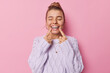 Leinwandbild Motiv Young glad woman points at toothy smile shows perfect white teeth being in good mood keeps eyes closed dressed in casual knitted jumper isolated over pink background. Positive emotions concept
