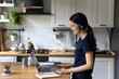 © fizkes - Side view woman standing in domestic cozy kitchen wear headphones having distance communication, looks at laptop use video call app, studying working remotely at home. Virtual meeting, e-learn concept