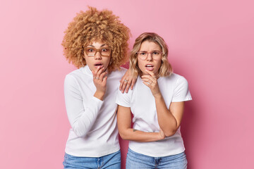 Wall Mural - Studio shot of shocked young women stare in amazement cannot believe their eyes hold breath from surprisement dressed in casual white t shirts isolated over pink background. Human reactions concept