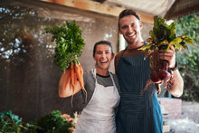 Pick Your Favorite. Portrait Of A Happy Young Couple Posing Together Holding Bunches Of Freshly Picked Carrots And Beetroot At Their Farm.