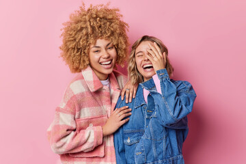 Wall Mural - Happy overjoyed young women laugh gladfully hear something very funny dressed in fashionable clothes stand closely to each other isolated over pink background. Friendship and emotions concept