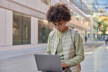 Outdoor shot of curly female student with curly hair dressed in casual clothes uses modern laptop prepares course work or new project poses in city. Female entrepreneur works remotely outside