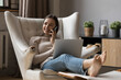 © fizkes - Positive busy young entrepreneur woman resting in armchair, working from home using laptop computer, talking on cellphone. Freelance worker, remote employee giving consultation on phone. Multitasking