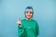 Positive hipster girl with colored hair stands on a blue background, shows his fingers a heart gesture and looks at the camera with a smile