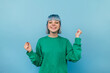 © bodnarphoto - Joyful hipster girl with blue hair stands on a blue background with a happy face raises her hands up and smiles, wears a green sweatshirt