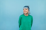 Fototapeta Uliczki - Positive lady with colored hair and in a green sweater stands on a blue background and looks away and laughs.