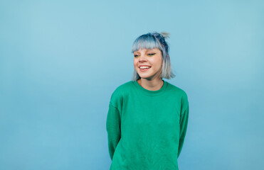 Positive lady with colored hair and in a green sweater stands on a blue background and looks away and laughs.