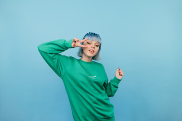 Wall Mural - Beautiful girl in a green sweatshirt and with colored hair poses for the camera on a blue background, shows a gesture of peace and laughs