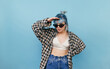 © bodnarphoto - Beautiful hipster girl with blue hair and sunglasses stands dressed in light summer clothes on a blue background and looks away.