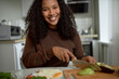 © Anatoliy Karlyuk - Smiling black woman cutting vegetables sitting at table in front of plate with fried eggs, wooden board, slicing peeled avocado against modern kitchen interior, wearing brown sweater