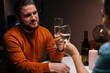 © dikushin - Close-up of happy young couple clinking glasses of white wine sitting at table with candles in dark cozy room. Love couple celebrating anniversary, Valentines day, enjoying romantic date.