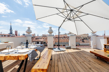 Poster - Terrace with large white fabric umbrella with wooden floors, solid wood dining table with benches and matching side table on an urban terrace of a vintage penthouse