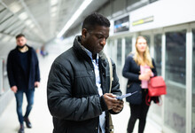 Portrait Of African American Male Passenger Waiting For Train On Subway Platform And Reading Timetable In Phone