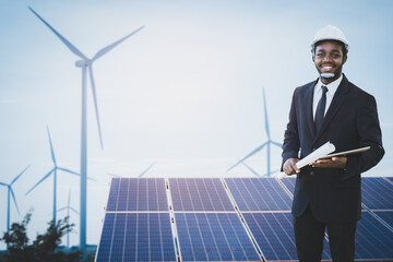 African engineer manager stand holding tablet and blueprint front solar photovoltaic and wind turbines generating electricity power station. Concept of sustainability development by alternative energy