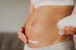 Pregnant woman putting cream on first trimester belly for stretch marks skin care prevention. Moisturizing dry skin during winter