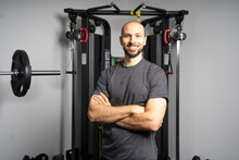 Smiling Fitness Instructor Standing With Arms Crossed In Front Of Exercise Equipment At Gym