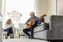 Senior Woman Looking At Man Playing Classical Guitar Sitting On Sofa At Home