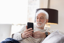 Senior Man Using Smart Phone Sitting On Sofa At Hotel Apartment