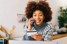 Happy Young Woman Holding Credit Card Talking On Mobile Phone At Home