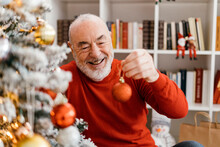 Happy Senior Man Holding Christmas Bauble Sitting In Living Room At Home