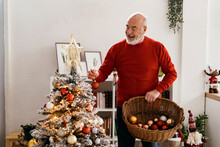 Senior With Basket Of Christmas Baubles Decorating Tree At Home