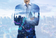 Attractive Young European Businessman With Folded Arms Standing On Abstract Blue City Background With Index, Candlestick Forex Chart And Mock Up Place. 