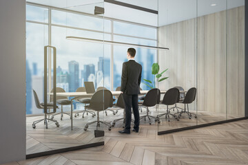 Wall Mural - Attractive european businessman standing in modern concrete and wooden, glass conference room interior with window and blurry city view, furniture and equipment.