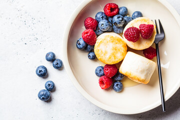 Canvas Print - Cottage cheese pancakes with raspberries, blueberries and syrup in white bowl, gray background. Russian cuisine concept.