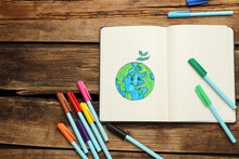 Happy Earth Day. Notebook With Drawing Of Planet And Markers On Wooden Table, Flat Lay