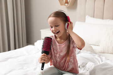 Wall Mural - Cute little girl in headphones with hairbrush singing on bed at home