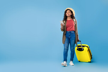 Young black woman with suitcase holding smartphone, booking hotel or vacation online, using web travel agency app