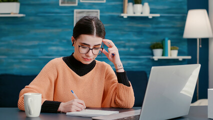 Wall Mural - University student attending online class on videoconference, using laptop to talk to teacher on remote video call meeting. Woman taking notes and having conversation on teleconference.